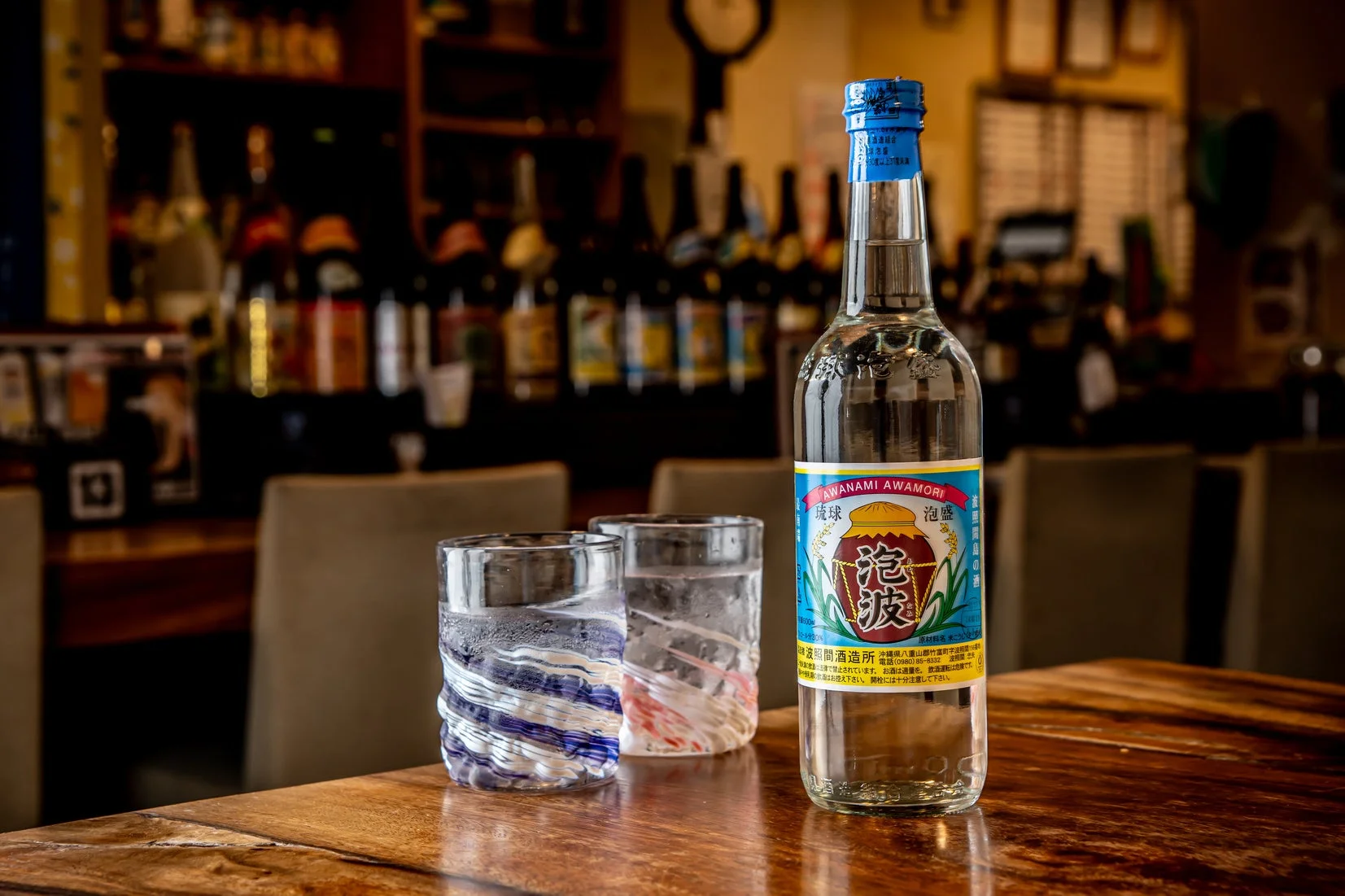 木製のテーブルに沖縄の伝統的なお酒である泡盛「泡波」のボトルと、水が入った二つの琉球ガラスのグラスが置かれています。