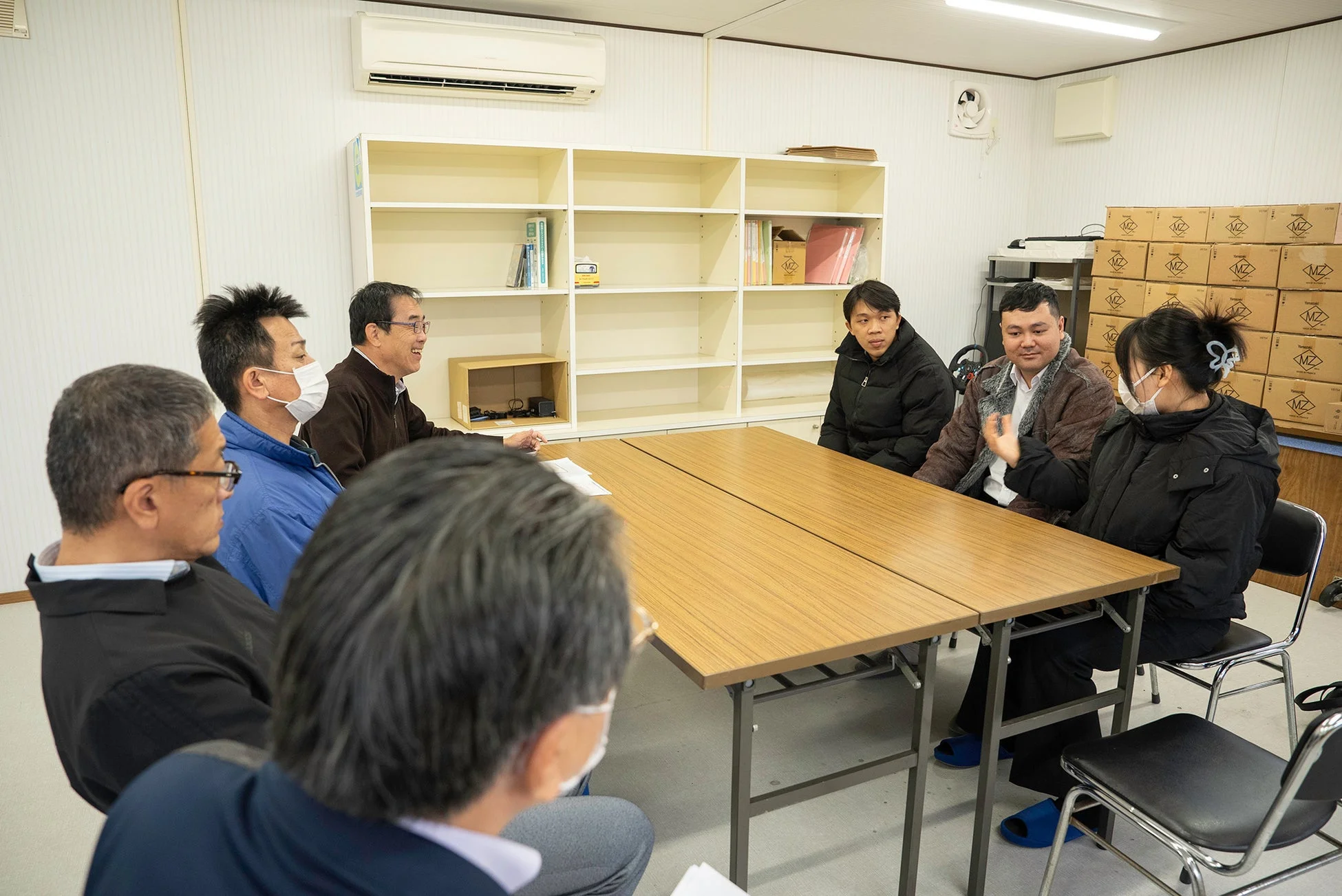 室内で複数の人々がテーブルを囲んで話し合っています。マスクを着用している人もおり、真剣な会議や打ち合わせが行われている様子です。