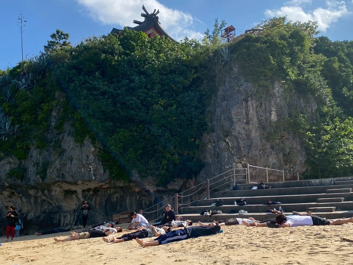 晴れた日、砂浜で人々が横たわりリラックスしている。背景には緑に覆われた崖があり、その上には伝統的な建物が見える。