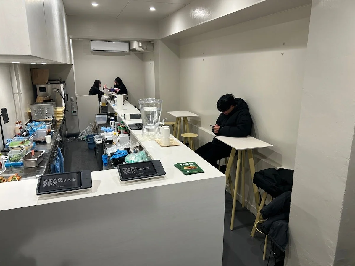 カジュアルな雰囲気の飲食店内部。客がスマートフォンを操作し、カウンターには「お会計はこちらから」の表示やバインミーのパンフレットが見える。