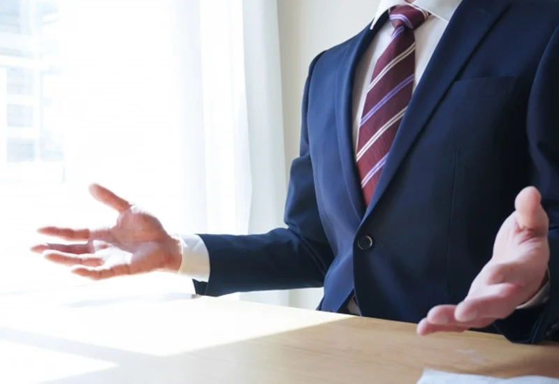 スーツを着た人物が両手を広げて何かを説明している