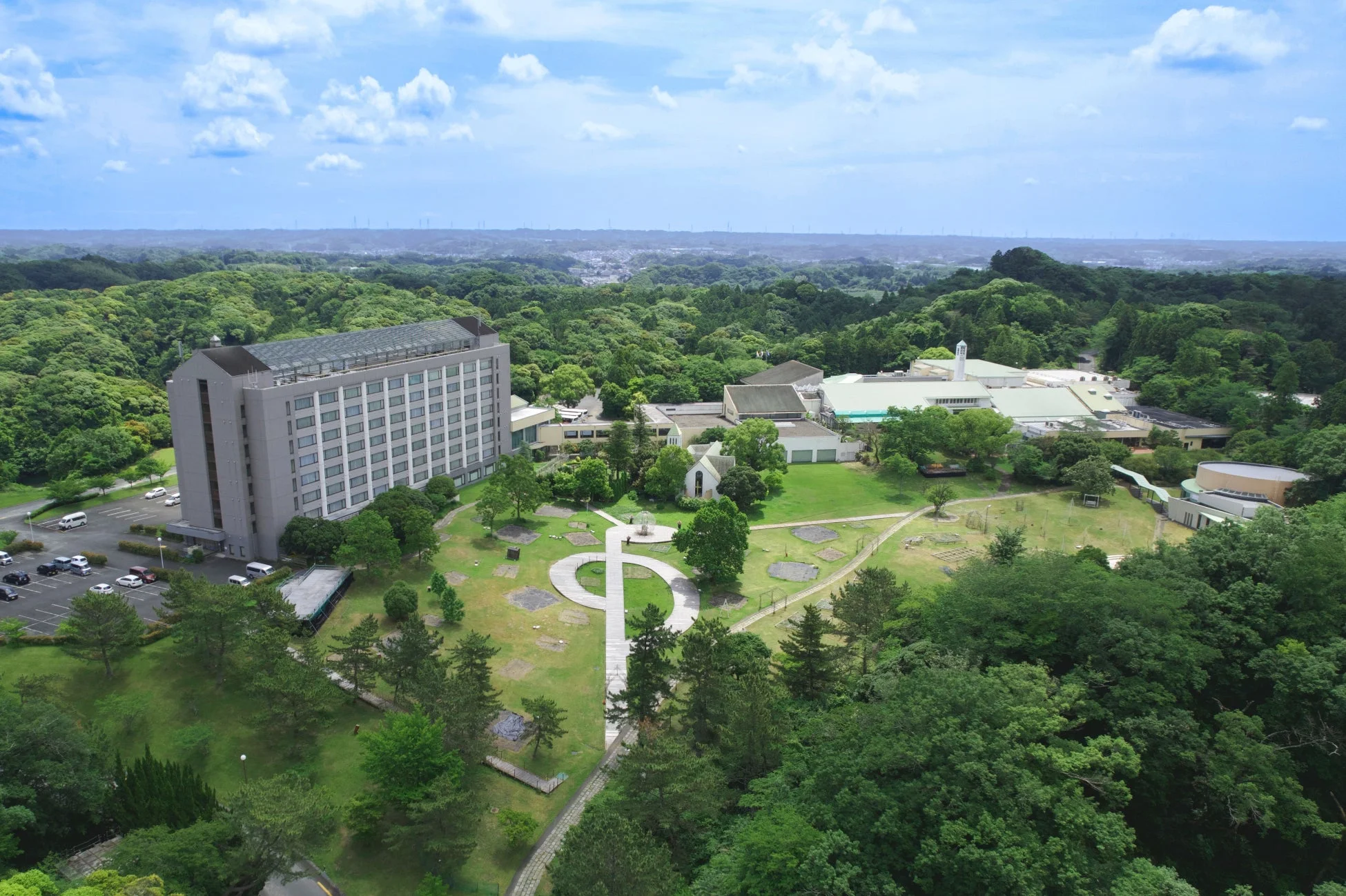 広大な緑に囲まれた敷地に、高層ビルや複数の建物が点在する景観。青空の下、整備された庭園や駐車場も見えます。