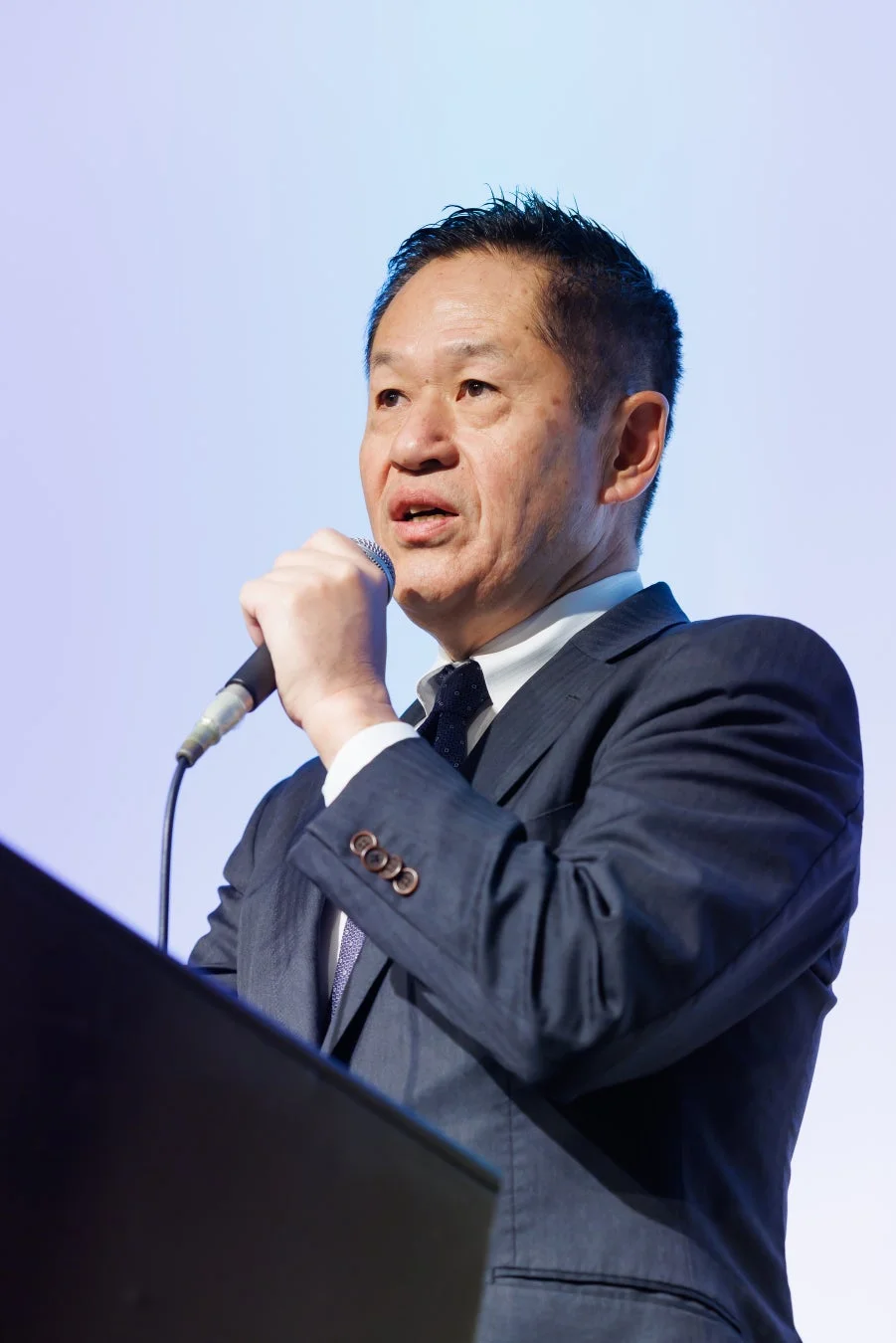 スーツを着用したアジア人男性が、マイクを手に演台の前で話している様子です。会議や講演会でのスピーチやプレゼンテーションを行っている場面と見られます。