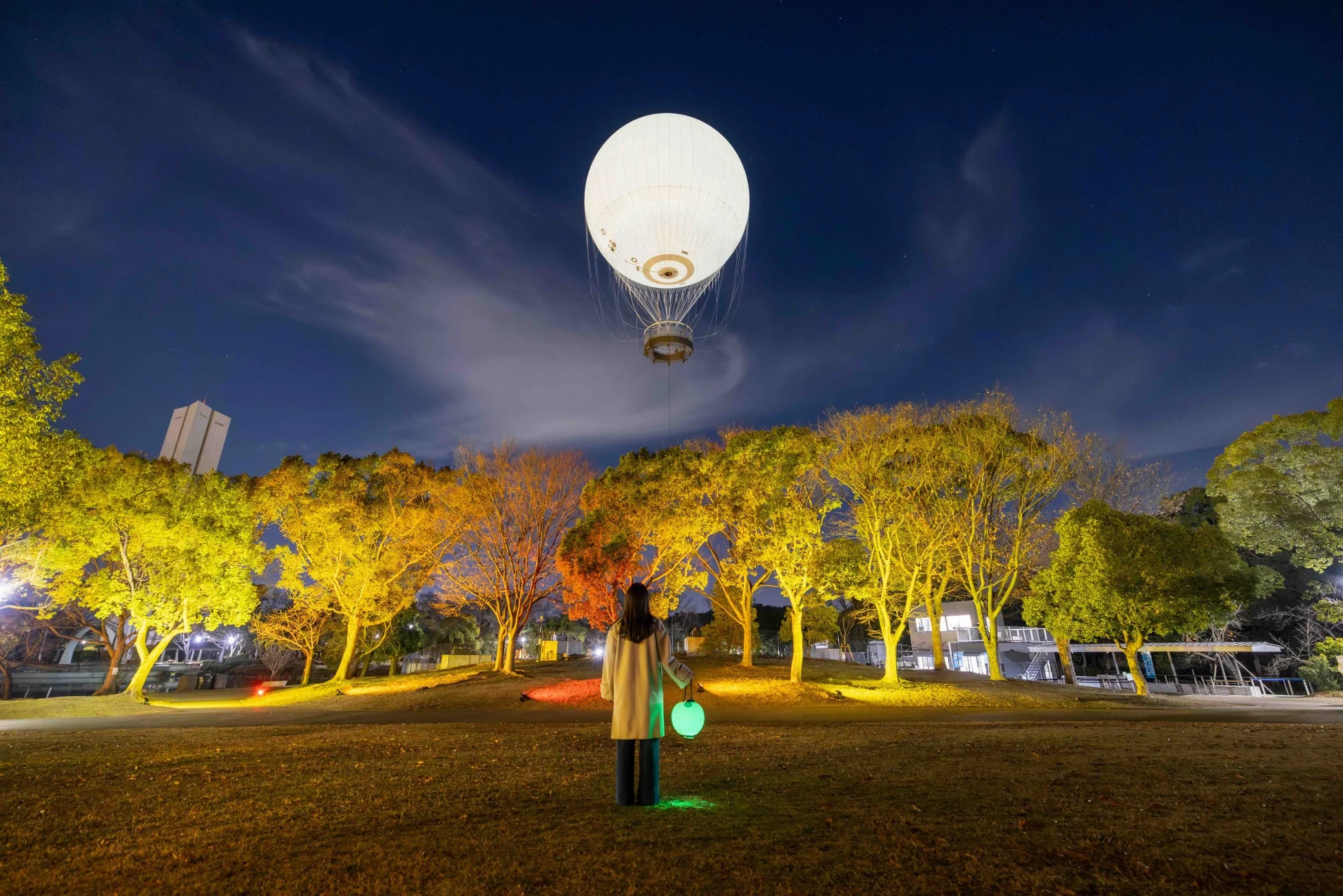 白く輝く熱気球とライトアップされた木々を背景に、緑のランタンを持つ人物が立つ夜の公園