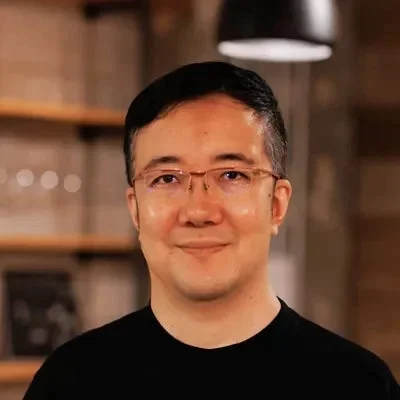 眼鏡をかけたアジア系の男性が黒いTシャツを着て、穏やかな笑顔を浮かべているポートレート写真