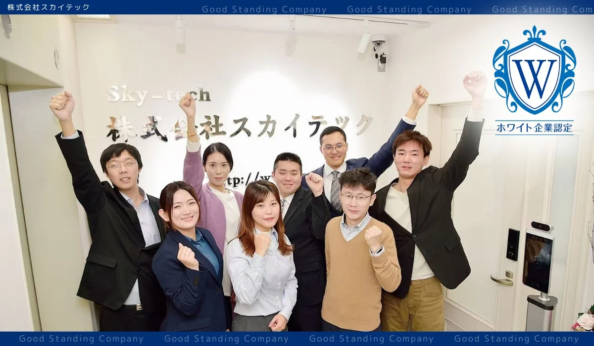 株式会社スカイテックの社員と思われる男女8名が、オフィス内で拳を突き上げるポーズを取り、笑顔で集合写真に収まっています。壁には会社名と「ホワイト企業認定」のロゴが見え、活気ある職場の雰囲気が伝わります。