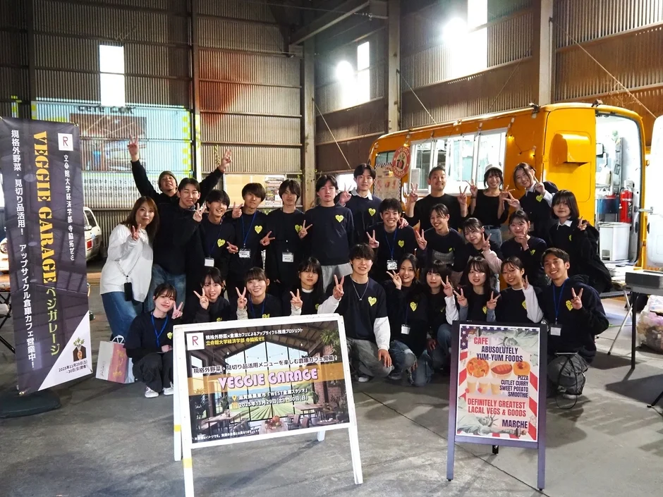 立命館大学の学生グループが、フードトラックのある倉庫のような空間で、規格外野菜を活用した食糧カフェ「VEGGIE GARAGE」のプロジェクトをPRしている集合写真です。多くの学生が笑顔でピースサインをしており、活気あるイベントの様子がうかがえます。