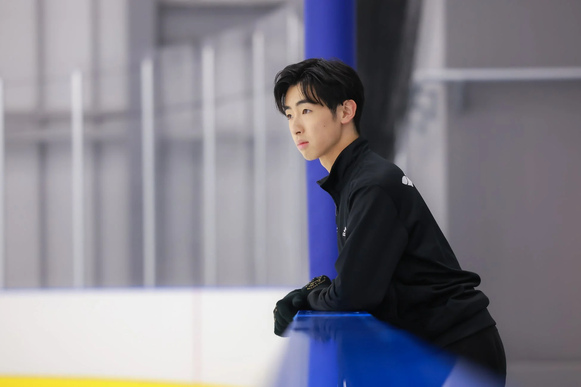 スケートリンクの青いフェンスに寄りかかり、思案げな表情で遠くを見つめる若い男性のフィギュアスケーター。