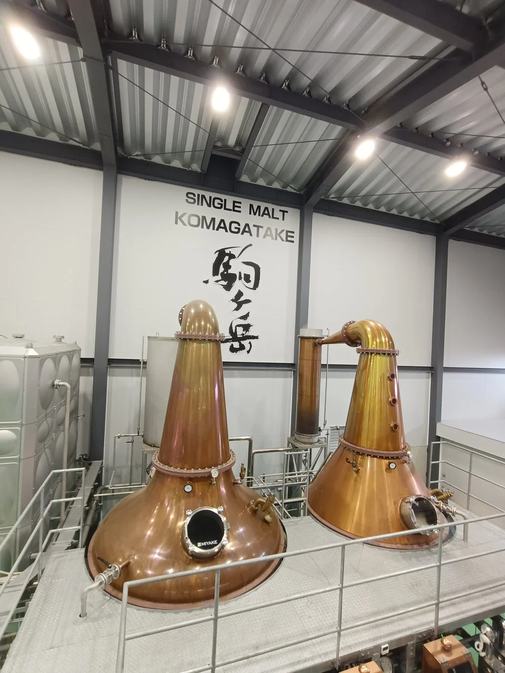ウイスキー蒸留所の内部で、美しい銅製のポットスチルが2基設置されている様子を捉えた画像です。壁には「SINGLE MALT KOMAGATAKE 駒ヶ岳」の文字があり、シングルモルトウイスキーの製造現場を示しています。