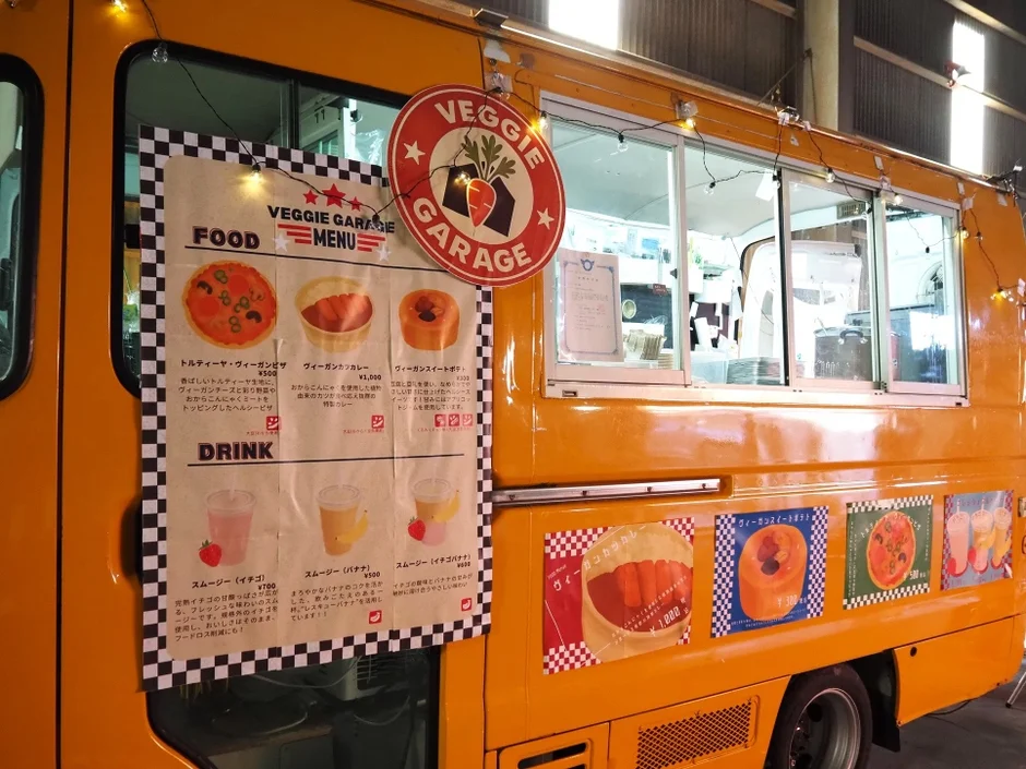 オレンジ色のフードトラック「VEGGIE GARAGE」のメニュー看板。ヴィーガンピザ、カレー、スイートポテト、各種スムージーなど、ヘルシーで環境に配慮したヴィーガン料理を提供している移動販売車の様子。