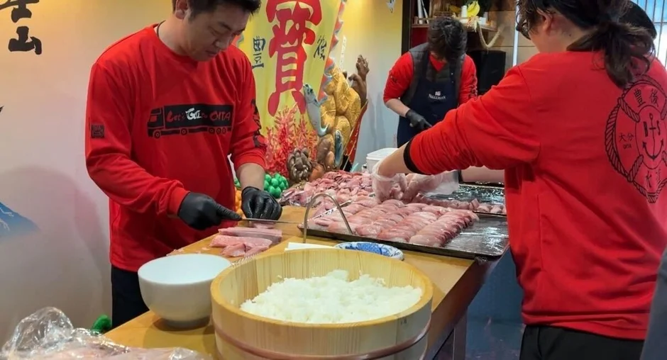 赤いシャツを着た人々が、大量の新鮮な魚を捌き、寿司や刺身の準備をしている様子。大きな木桶に入ったご飯もあり、大分県をテーマにしたイベントか市場での調理風景と見られる。