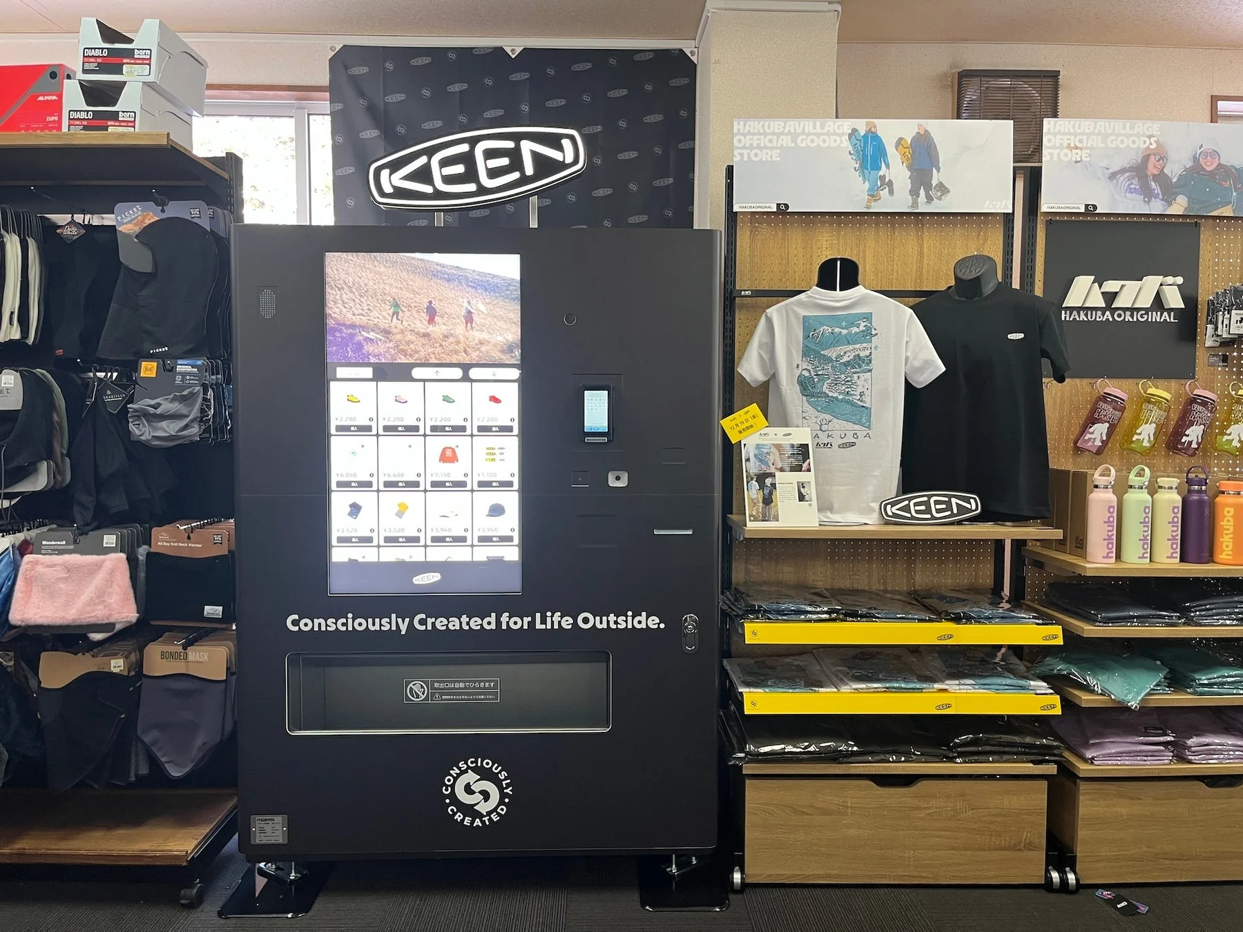 アウトドアブランドKEENの製品と白馬村のオリジナルグッズが並ぶ店舗の様子です。衣料品やボトルが陳列され、靴や小物も販売する自動販売機が設置されています。