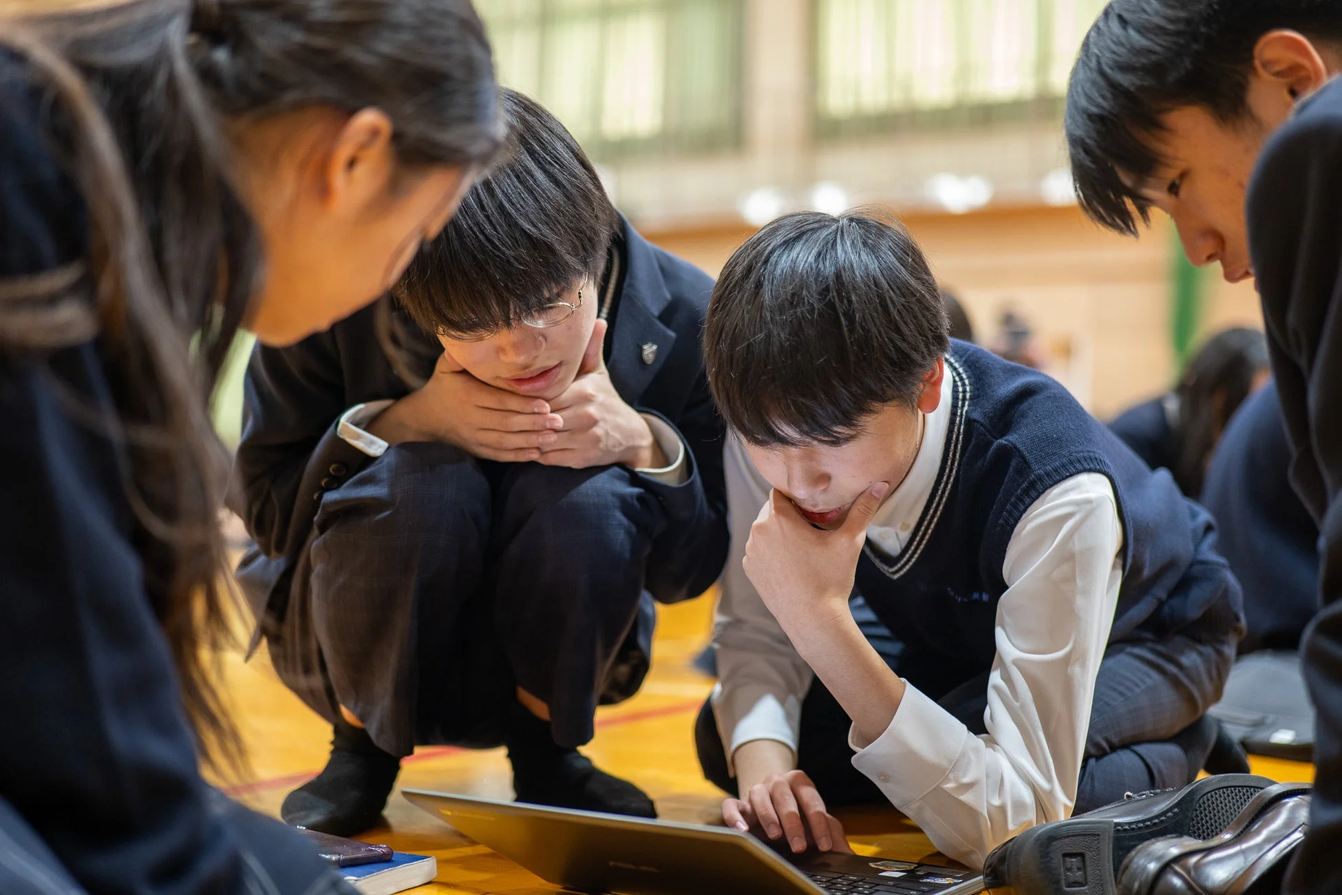 数人の生徒が体育館のような場所で、ノートパソコンを囲んで集中して話し合っています。制服を着ており、グループ学習や共同作業をしている様子が伺えます。