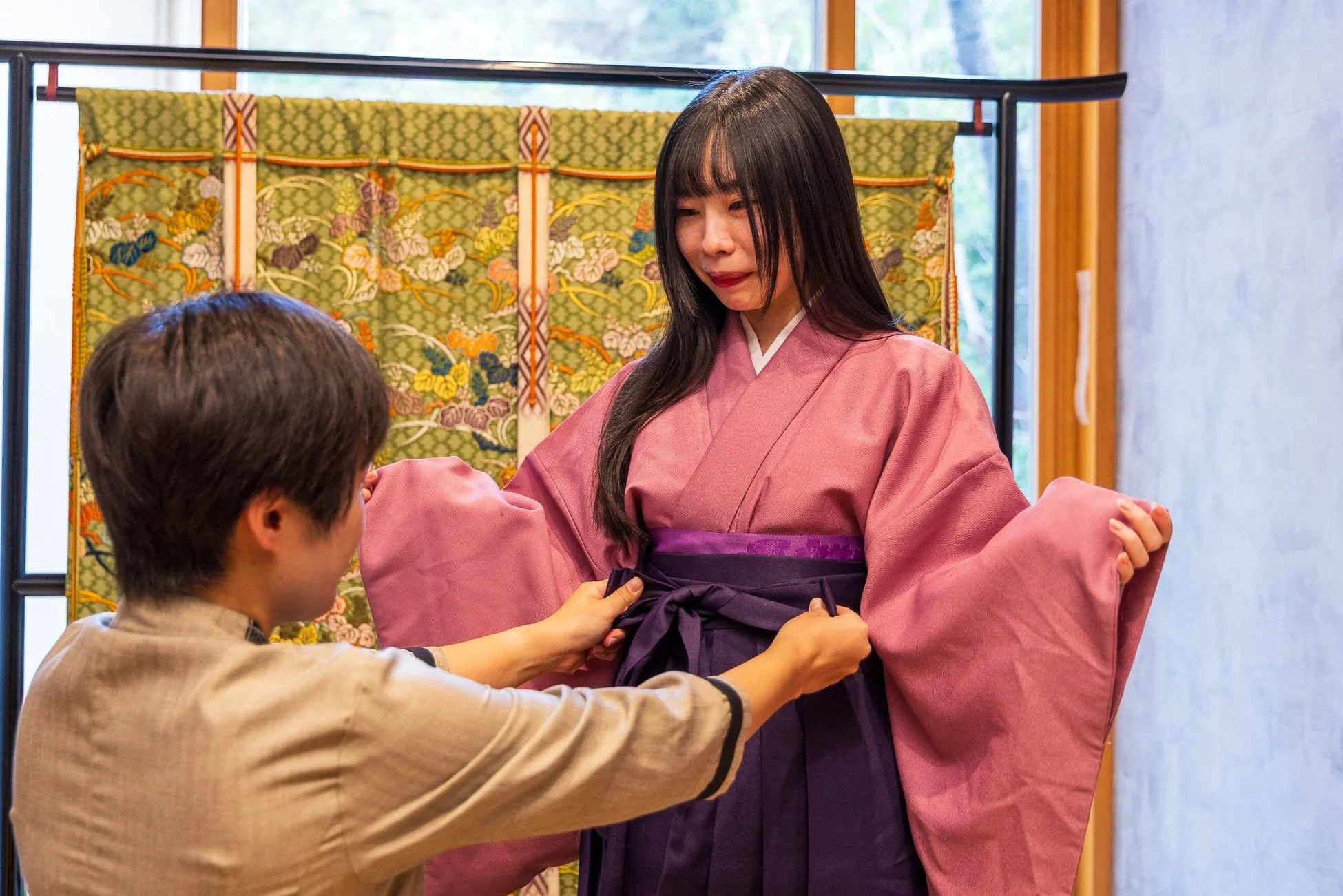 ピンクの着物と紫の袴を着用した女性が、着付けをしてもらっている様子