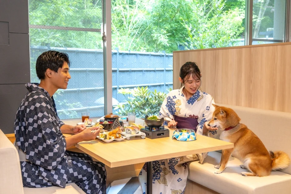 浴衣を着た男女が、柴犬と一緒に和やかな雰囲気の中で食事を楽しんでいる様子です。窓からは緑が見え、日本料理を囲んでリラックスしたひとときを過ごしています。ペット同伴可能な施設での食事風景を捉えた一枚です。