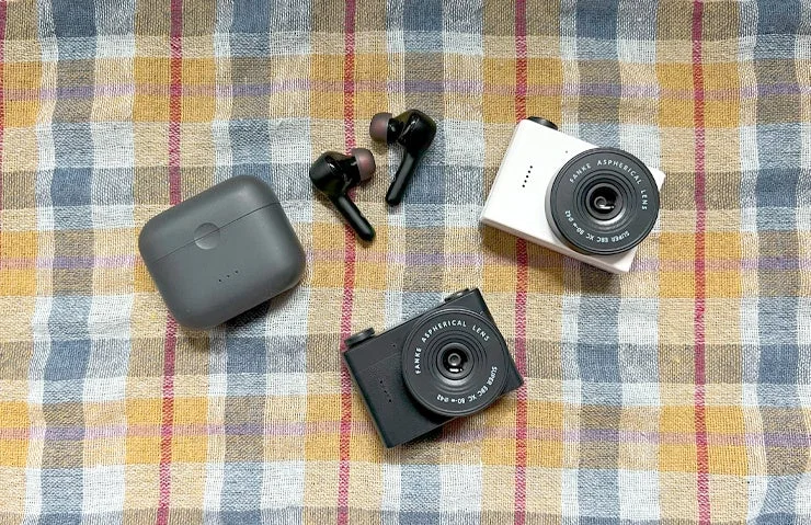 チェック柄の布の上に、白と黒のコンパクトカメラ2台、黒いワイヤレスイヤホンとその充電ケースが平置きされています