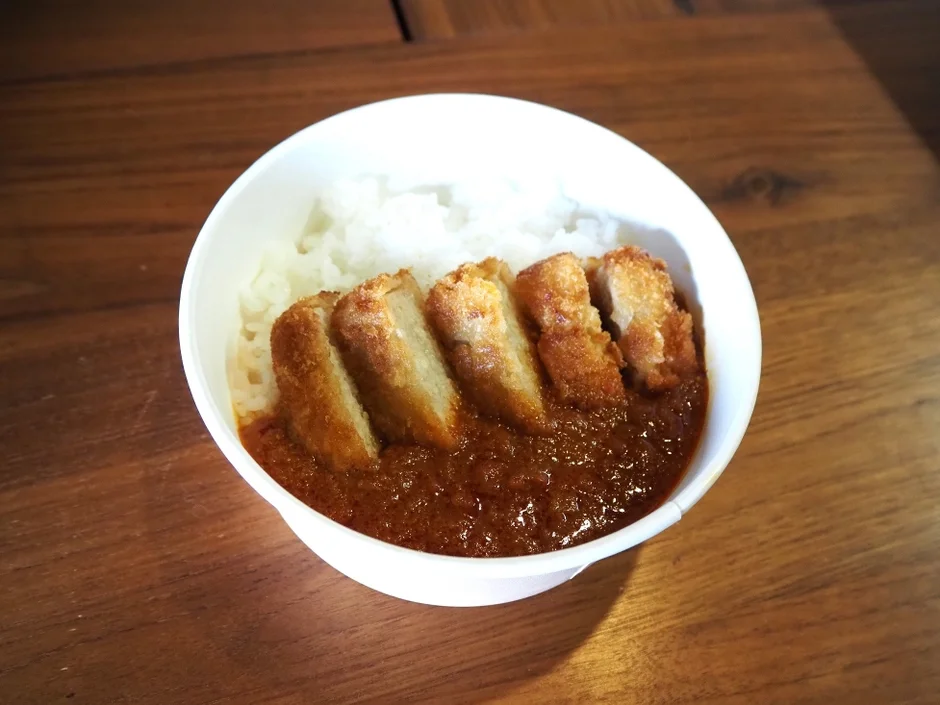 白い器に盛られたご飯の上に、揚げたてのカつが数切れ乗せられ、その周りに濃厚なカレールーがたっぷりとかけられています。木製のテーブルに置かれた、食欲をそそるカツカレーのクローズアップ写真です。