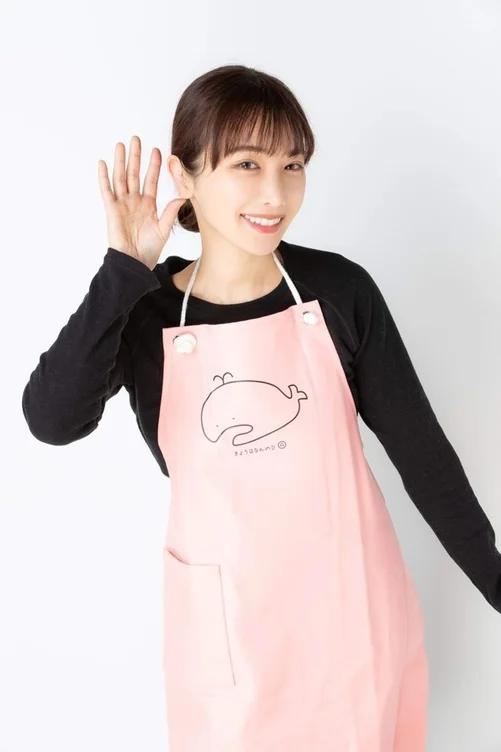 ピンク色のクジラのイラストが入ったエプロンを着用した女性が、笑顔で耳に手を当てて聞くポーズをとっています。明るく親しみやすい雰囲気です。