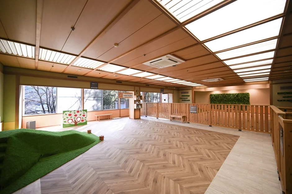 広々とした明るい屋内のキッズパークで、木目調の床と天井が特徴的です。人工芝の小山やリンゴの絵が描かれたボードがあり、子供たちが楽しく遊べるよう設計されています。窓からは外の景色が見え、開放感があります。
