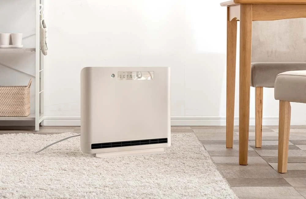 明るい部屋の白いラグの上に置かれたベージュ色の暖房器具または空気清浄機。右には木製のテーブルと椅子、左には棚が見え、全体的にシンプルで清潔感のあるインテリアです。