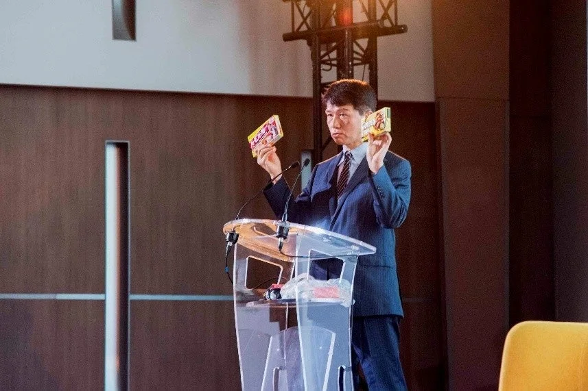 ポッキーとプリッツの箱を持ってプレゼンテーションを行う井上岳久学長