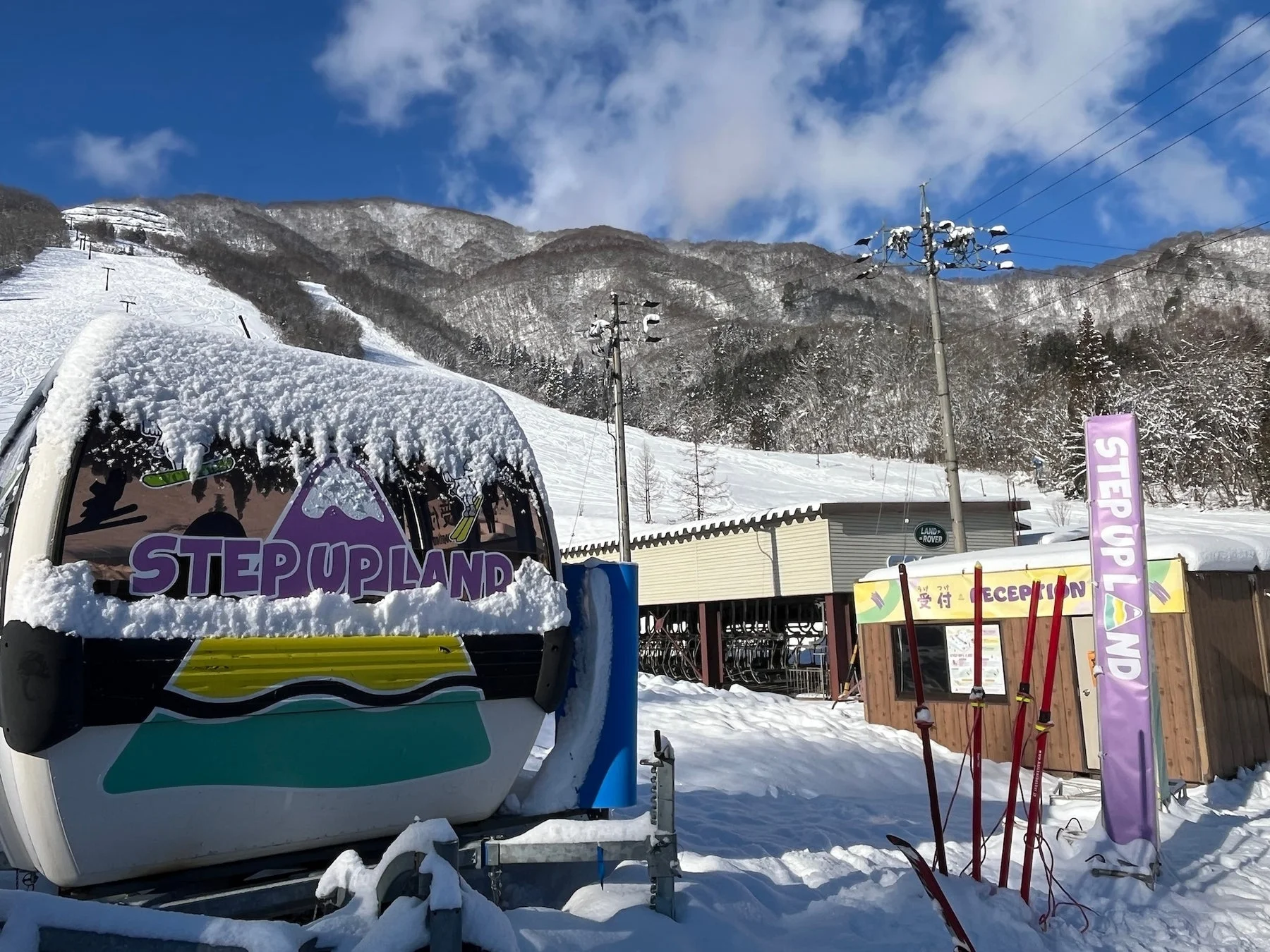 晴れた冬の日に雪が積もったスキー場の風景です。ゴンドラやスキーリフト、受付の建物が見え、遠くには雪山が広がっています。スキーシーズン中の活気ある様子が伝わります。