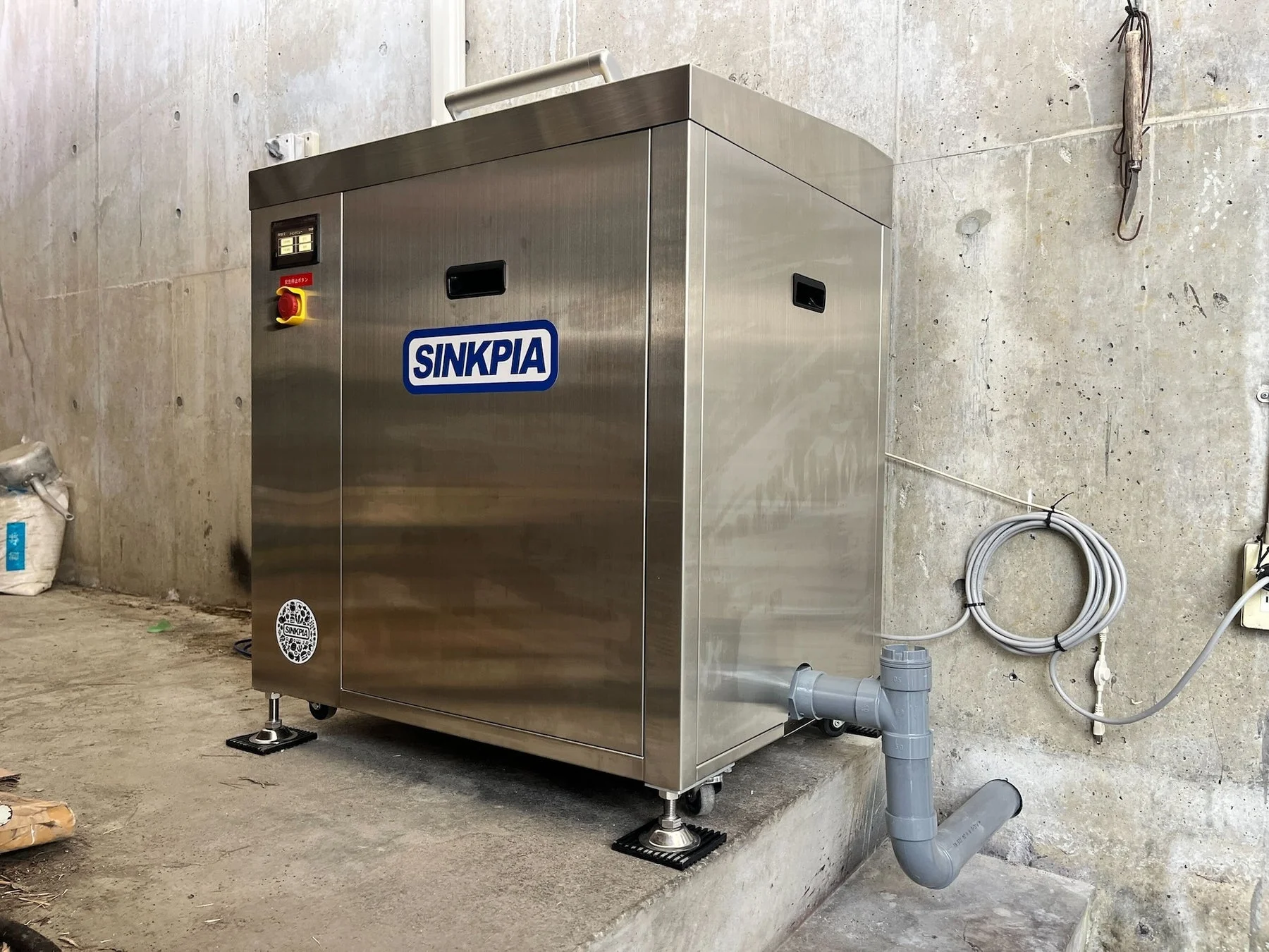 ステンレス製の業務用機械「SINKPIA」がコンクリートの壁と床の空間に設置されている写真。排水パイプや制御パネル、非常停止ボタンが見える。