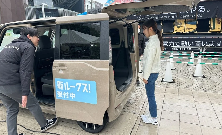 日産の新型車「ルークス」の展示イベントの様子。ベージュのルークスがドアを開けた状態で展示されており、男性と女性が車内を覗き込んでいる。背景には「電力で動いています」「SAKURA」と書かれたバナーが見える。