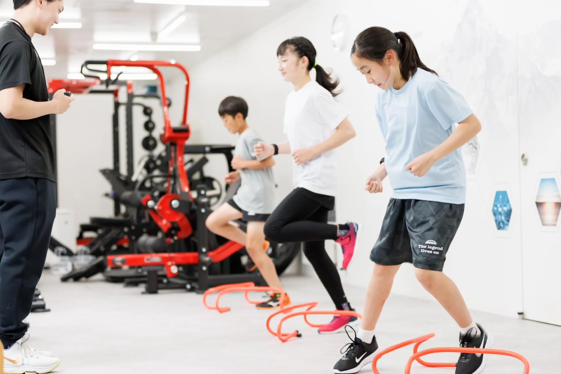 室内ジムで、コーチの指導のもと、子供たちがミニハードルを使ったアジリティトレーニングに励んでいる様子