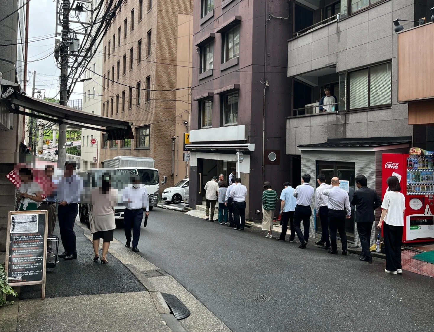 日本の街角で、多くの人々が飲食店と思われる建物の前に行列を作っている様子。