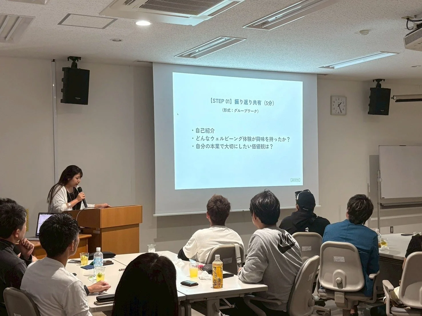 女性がマイクを持ってプレゼンテーションを行っており、参加者がスクリーンに映し出された「振り返り共有」のテーマを見ながら話を聞いている様子。