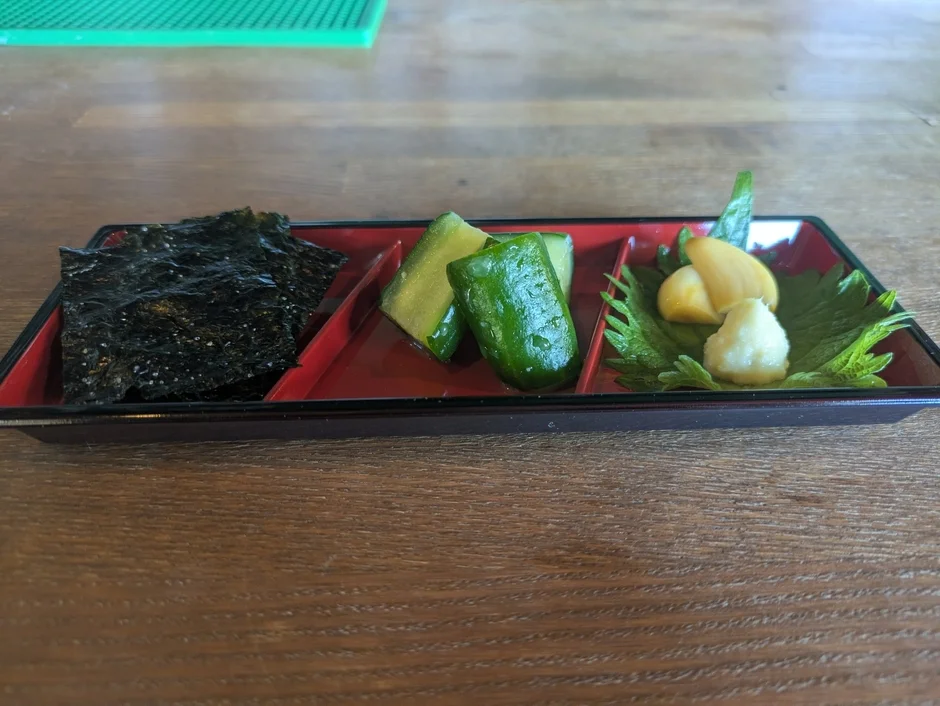木製のテーブルに置かれた黒いお盆に、海苔、きゅうりの漬物、そして薬味（おそらく生姜やニンニクの漬物とすりおろしたもの）が美しく盛り付けられています。日本の伝統的な軽食やおかずの一品です。