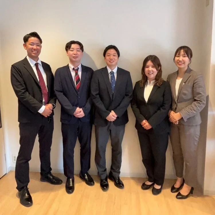 ビジネススーツを着用した5人の男女が、白い壁の前で笑顔で並んで立っている集合写真