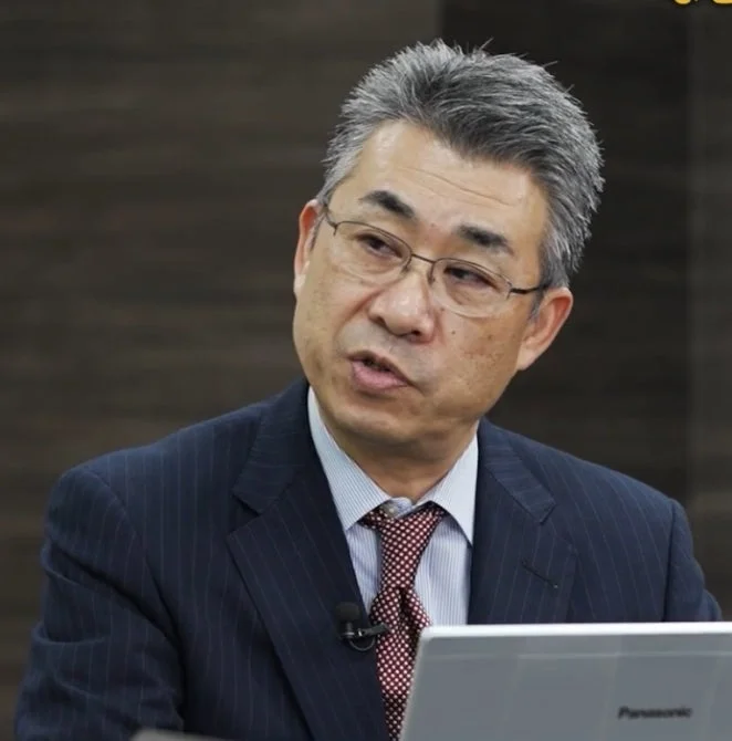 スーツを着た中年のアジア人男性が、マイクを付けた状態でラップトップの前で話している様子