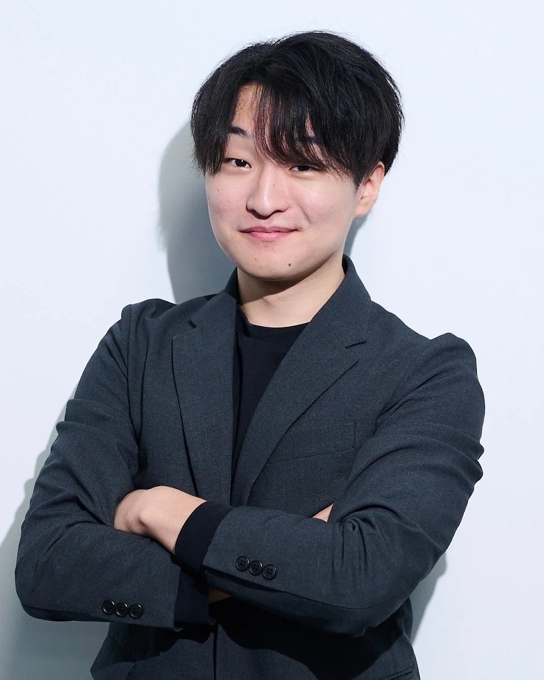 白い背景の前で、ダークグレーのスーツを着用し腕を組んで微笑むアジア人男性のポートレート