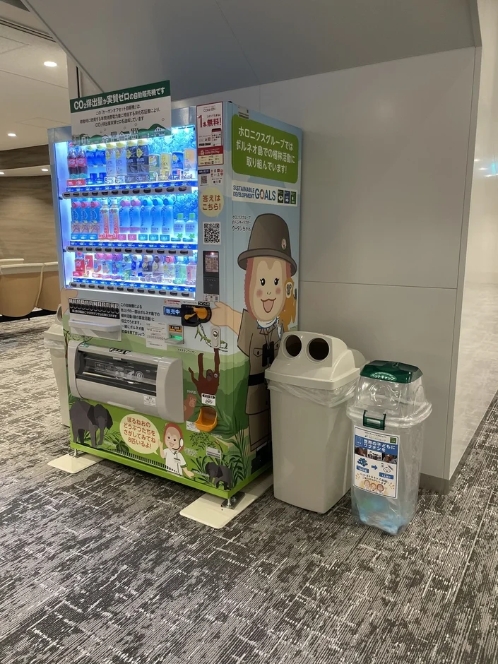 CO2排出量実質ゼロの自動販売機と、その隣に設置されたゴミ箱が写っています。自動販売機はボルネオ島の植林活動を支援し、ペットボトルキャップは世界の子供たちへのワクチン寄付に繋がる活動をアピールしています。
