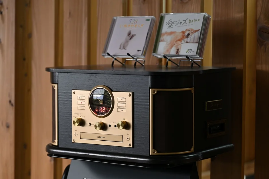 木目調の壁を背景に、レトロなデザインの黒とゴールドのステレオシステムが置かれています。上には犬のジャケットが特徴的なCD2枚が飾られ、音楽を楽しむための多機能オーディオ機器のようです。