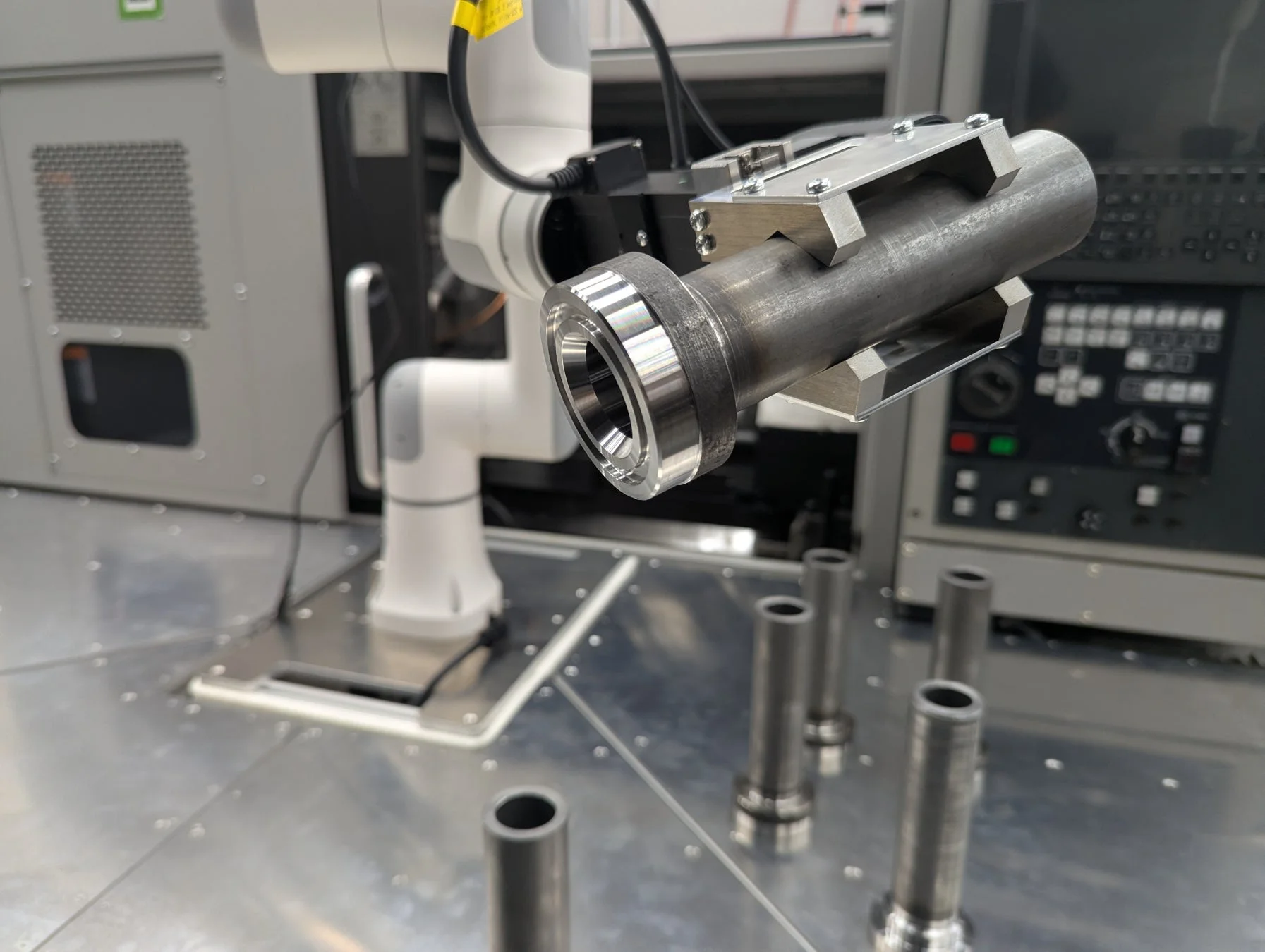 ロボットアームが金属製の円筒形部品を把持しており、自動化された製造工程の一部を示しています