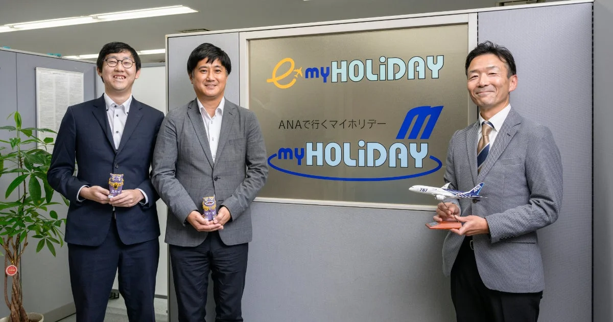 スカイツアーズの「myHOLIDAY」ロゴの前で笑顔で立つ男性たち