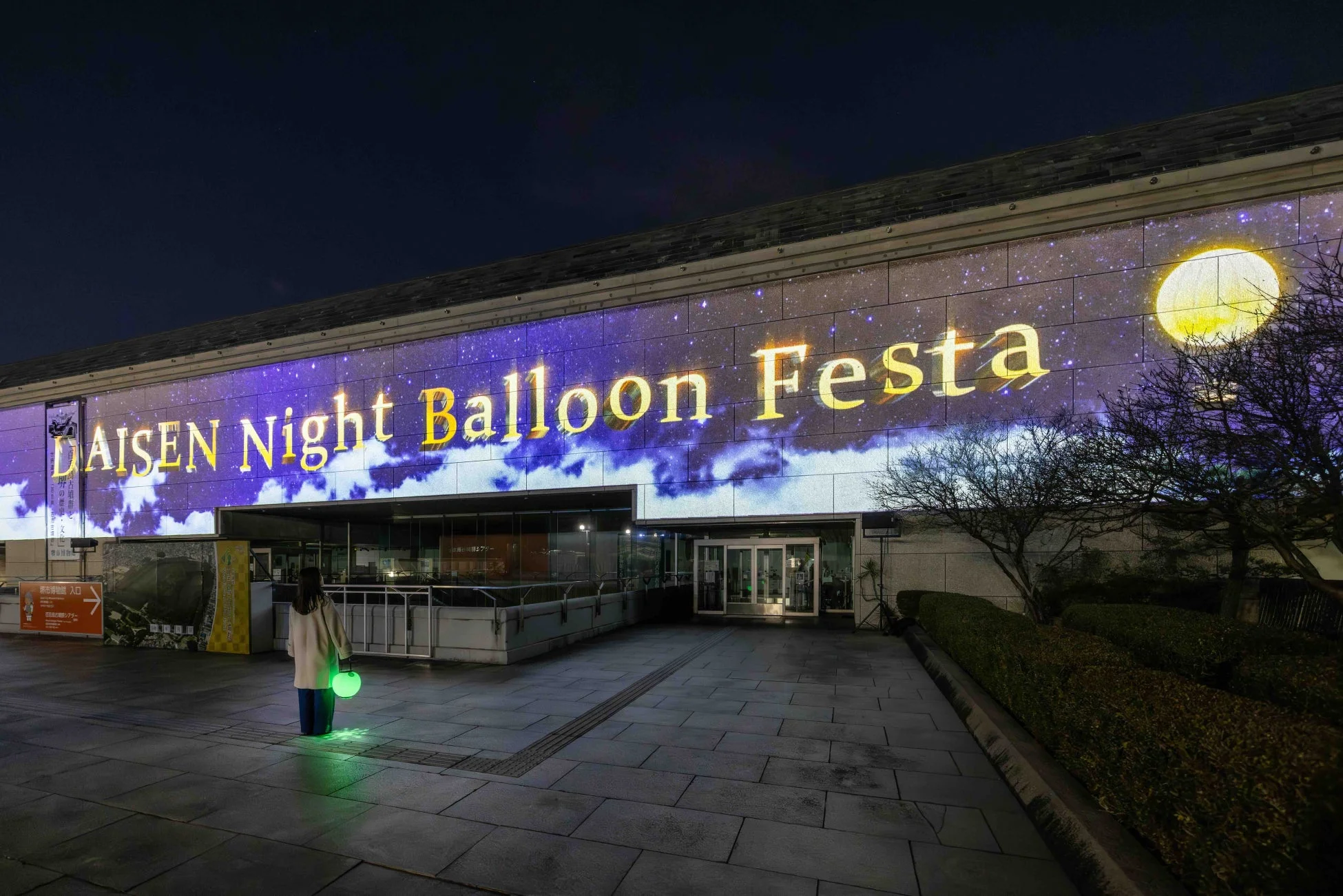 夜の建物にプロジェクションマッピングが映し出され、光る球体を持つ人が立つ幻想的な雰囲気