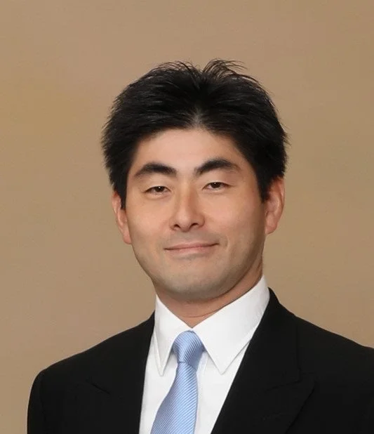 黒いスーツと水色のネクタイを着用したアジア人男性のポートレート写真です。彼はカメラに直接向き合い、穏やかな笑顔を浮かべています。真面目さと親しみやすさを兼ね備えた印象です。