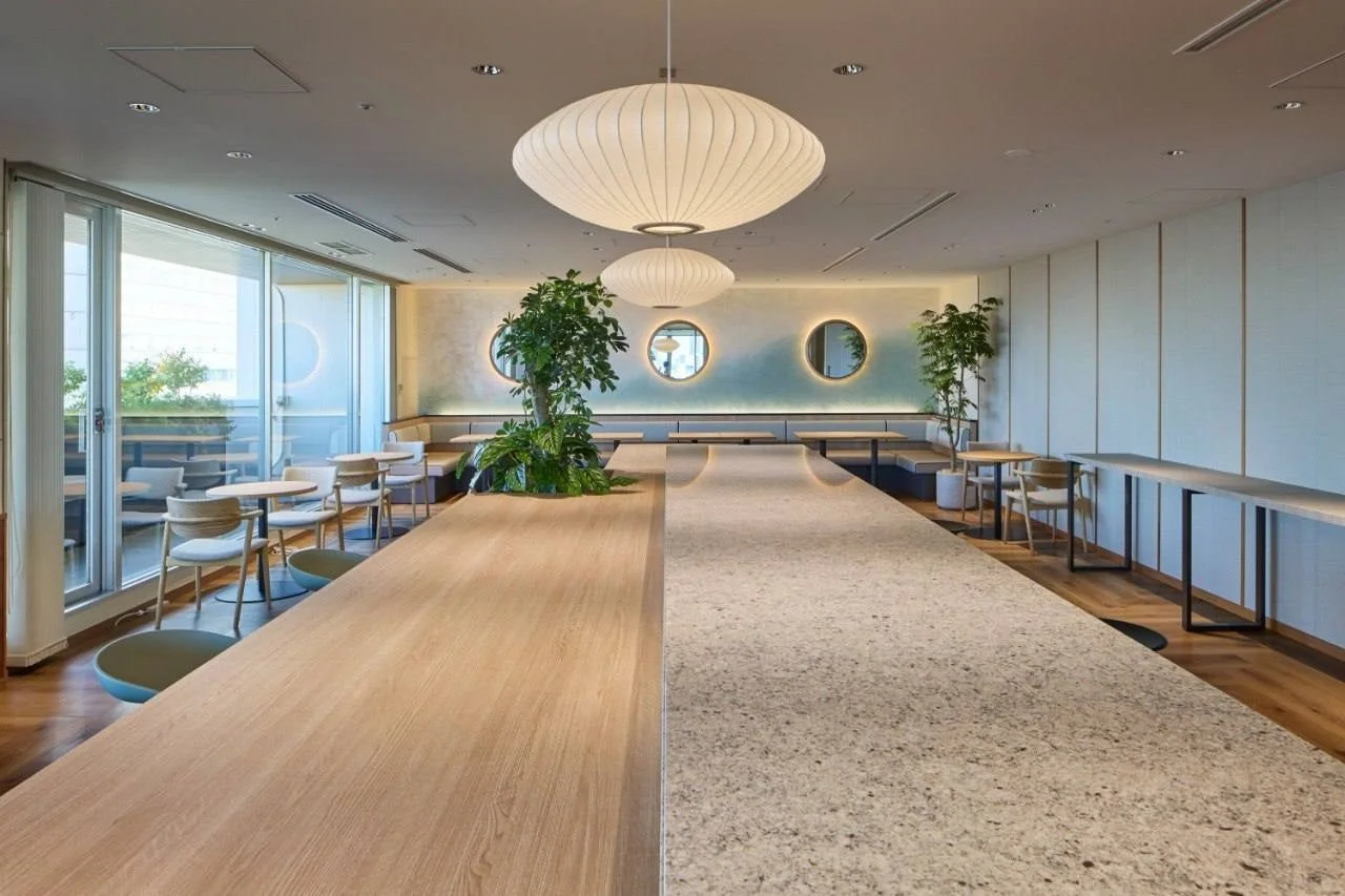モダンで広々とした明るい空間が特徴のラウンジやカフェ、オフィススペースです。大きな窓から自然光が差し込み、木目調のテーブルや植物、デザイン性の高い照明が配置され、リラックスできる雰囲気を醸し出しています。