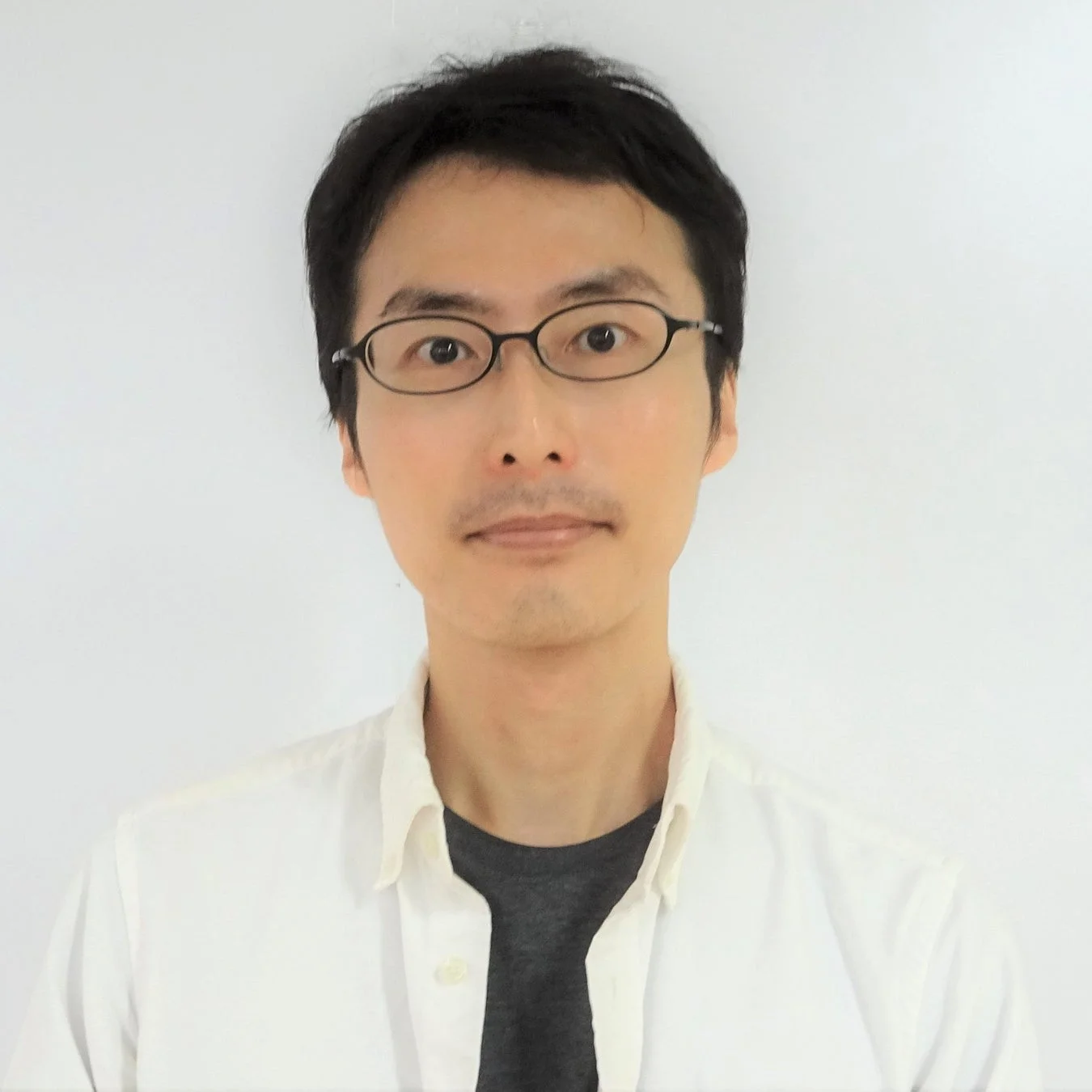 白い背景の前で、メガネをかけたアジア人男性が白いシャツを着てカメラを見つめている上半身のポートレート写真です。