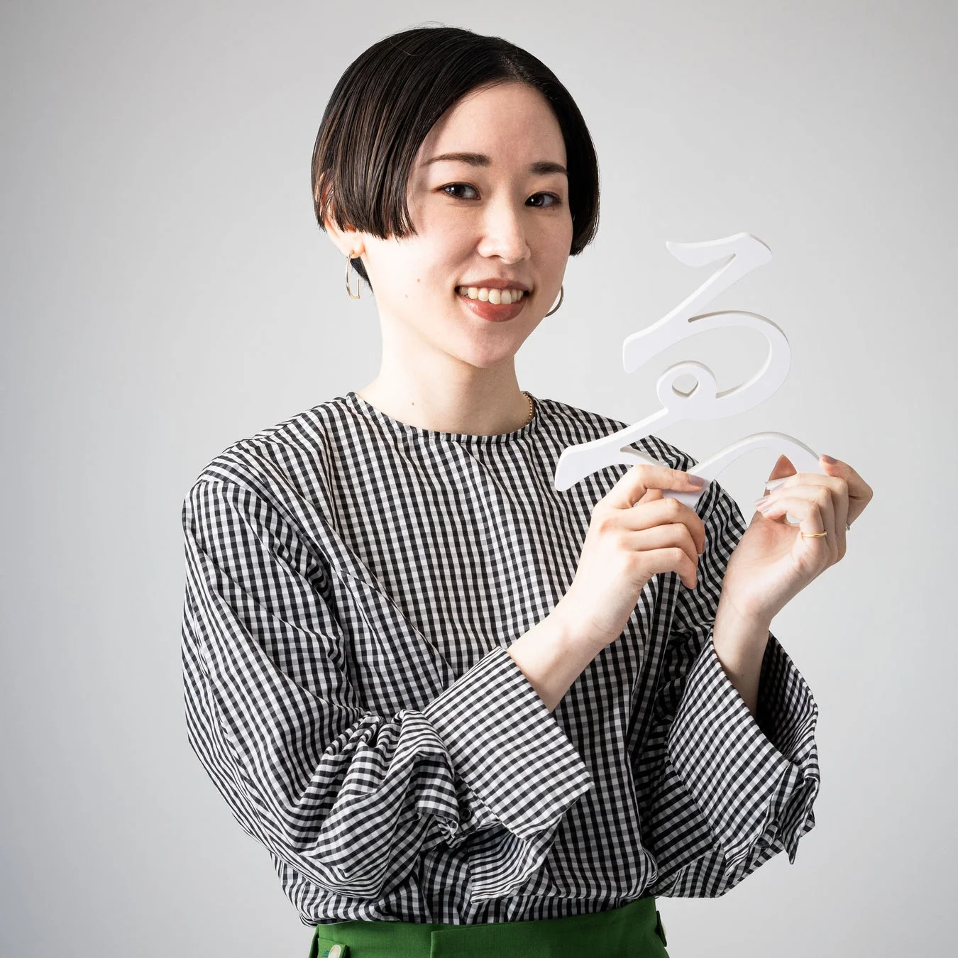 白い背景の前で、ギンガムチェックのブラウスと緑色のパンツを着用したショートヘアの女性が笑顔でポーズを取っています。彼女は白いひらがなの「る」の文字を両手で持っています。