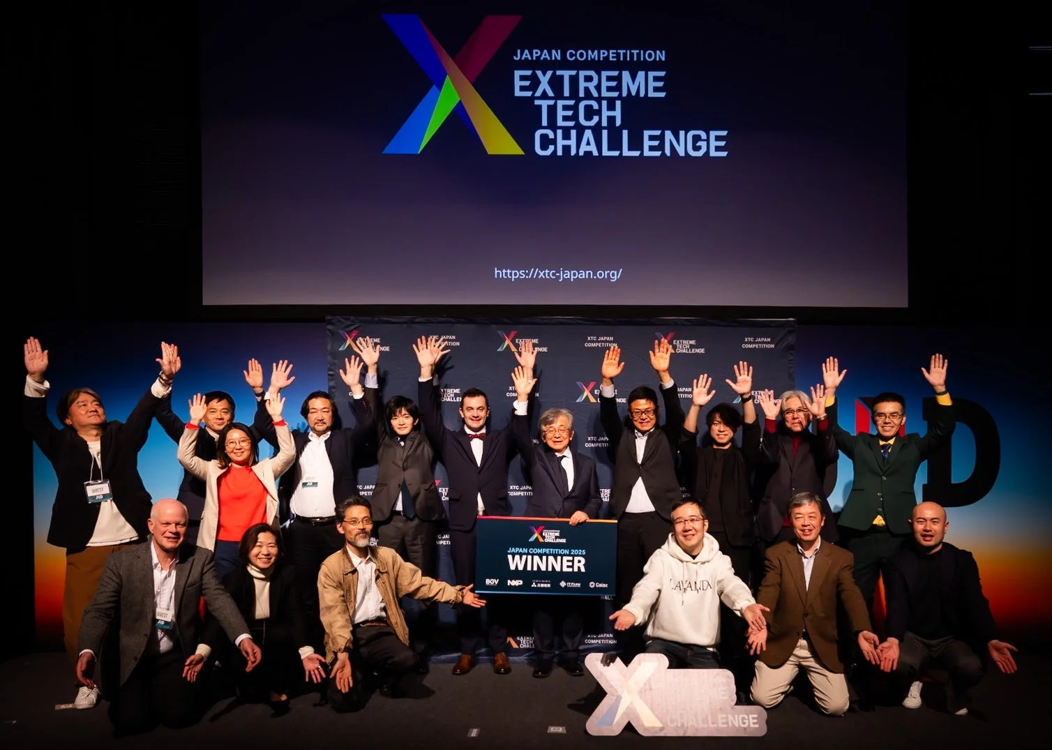 XTC Japan Competition 2025のイベントで、参加者や関係者が手を上げて祝っている集合写真