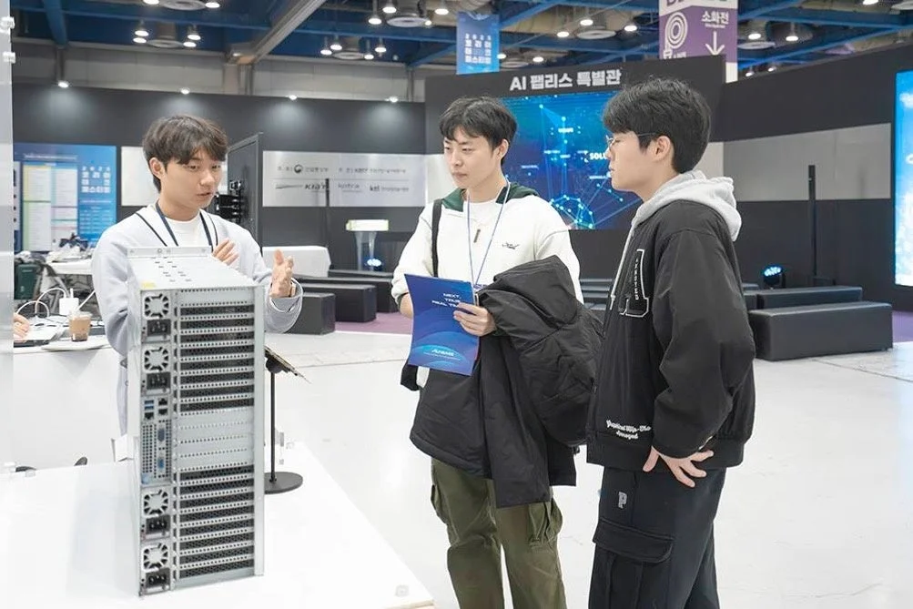 AI関連の展示会で、一人の男性が目の前のサーバーラックについて他の二人へ説明している様子。彼らは技術的な内容について話し合っているようだ。