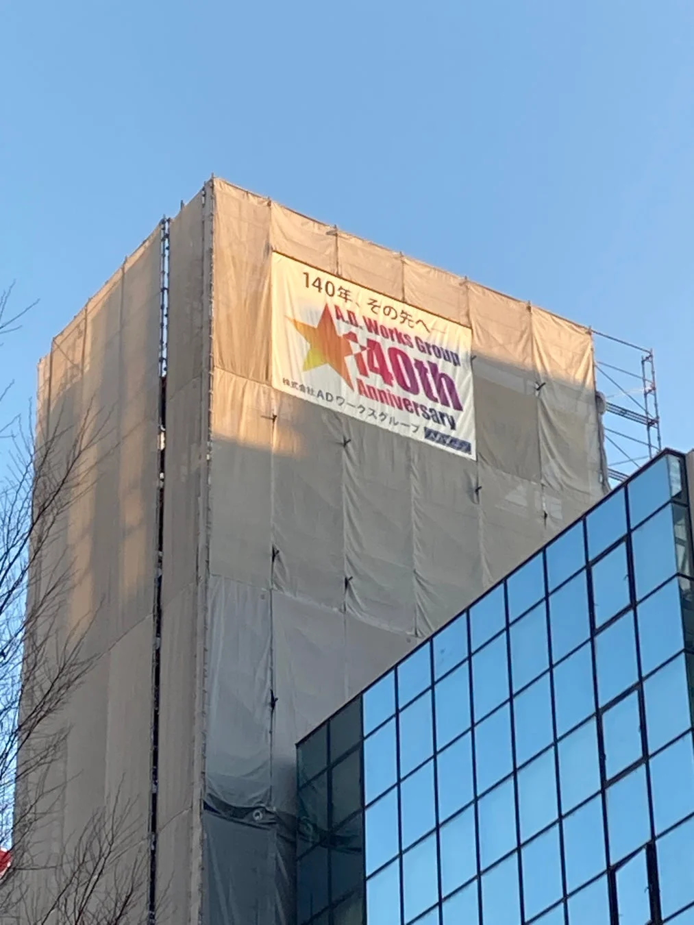 足場と白いシートで覆われた建設中の建物に、ADワークスグループの40周年を祝う大きなバナーが掲げられています。青空の下、隣接するガラス張りの近代的な建物も一部見えています。