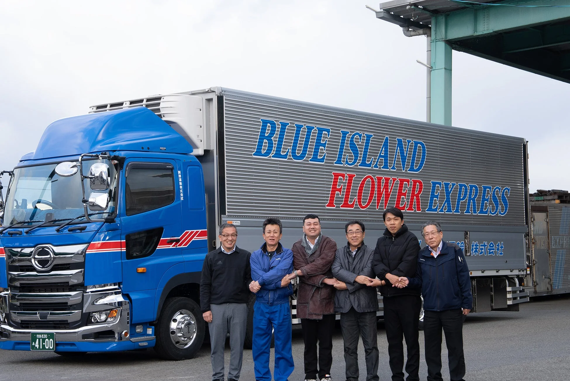 BLUE ISLAND FLOWER EXPRESSと書かれた青い日野の冷蔵トラックの前で、藍通運輸株式会社の男性社員6人が並んで立つ集合写真です。