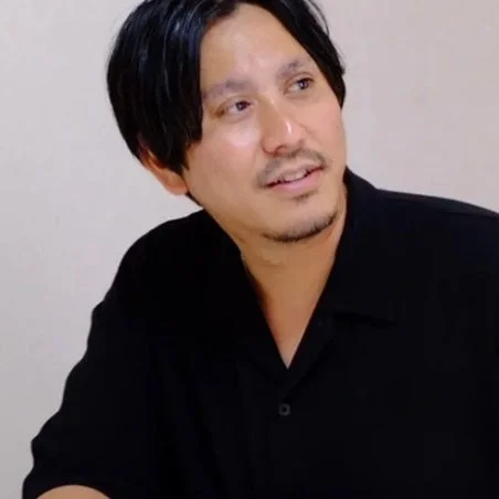 黒髪で髭と口ひげのある男性が黒いシャツを着ており、画面の右側を見ている