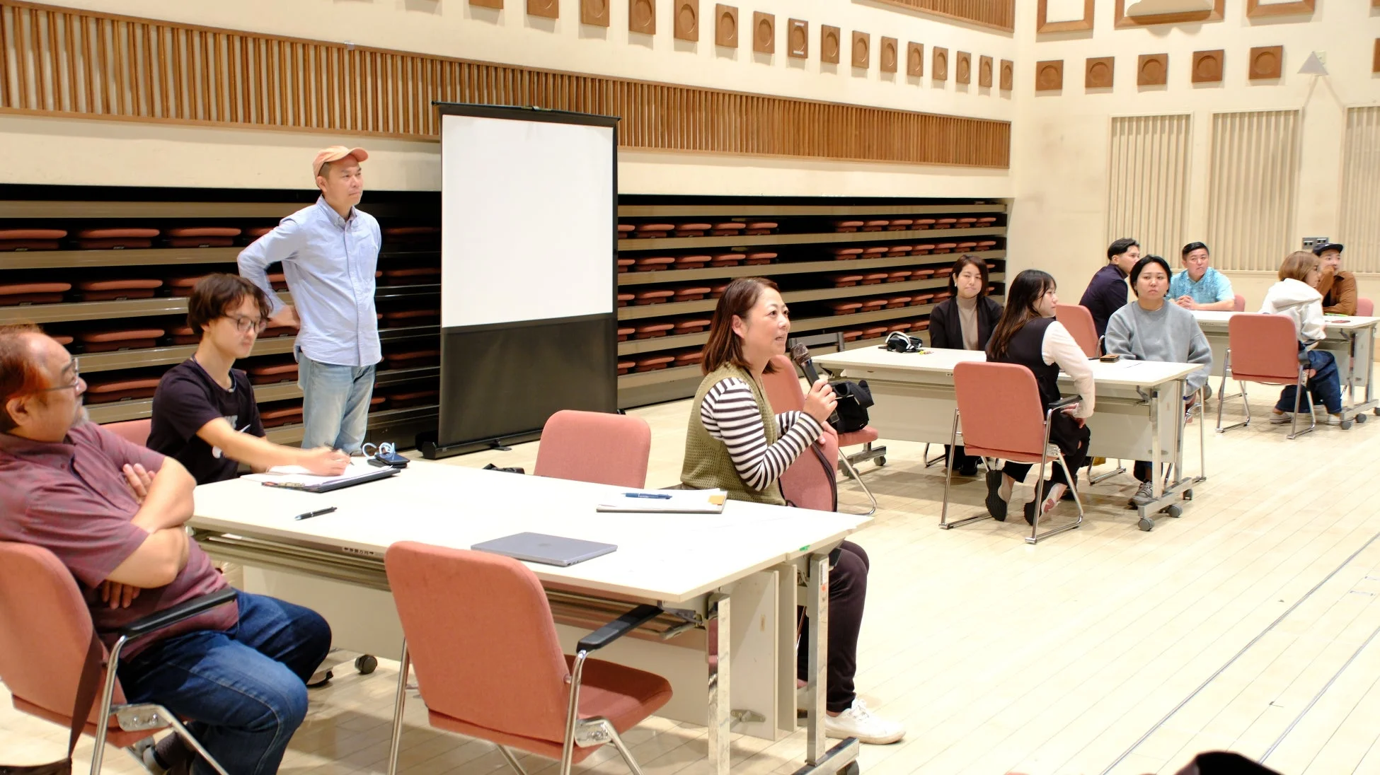 会議室のような場所で、複数の参加者がテーブルに着席し、一人の女性がマイクを持って話している様子